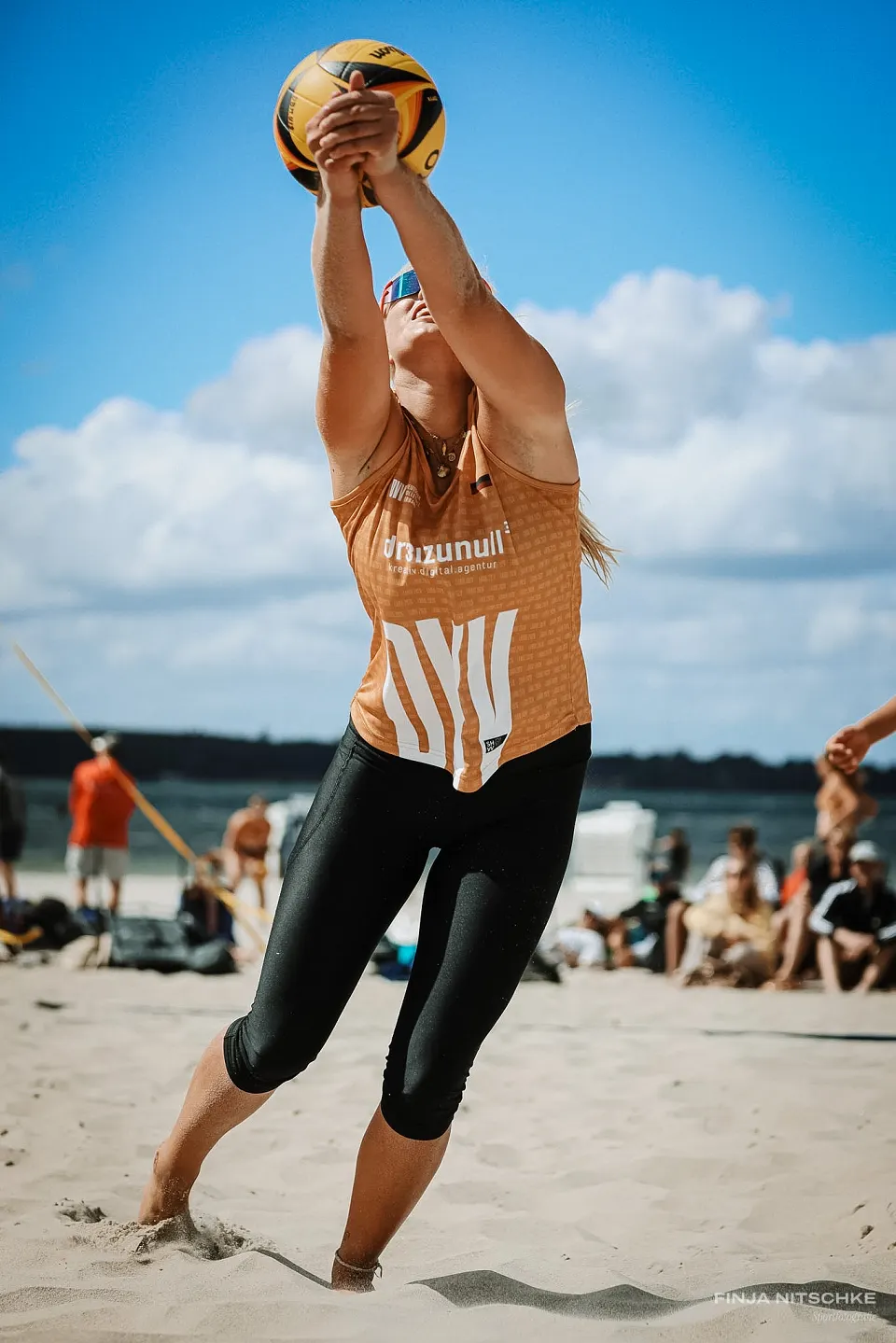 Beachvolleyballerin streckt sich mit beiden Armen nach dem Ball unter blauem Himmel