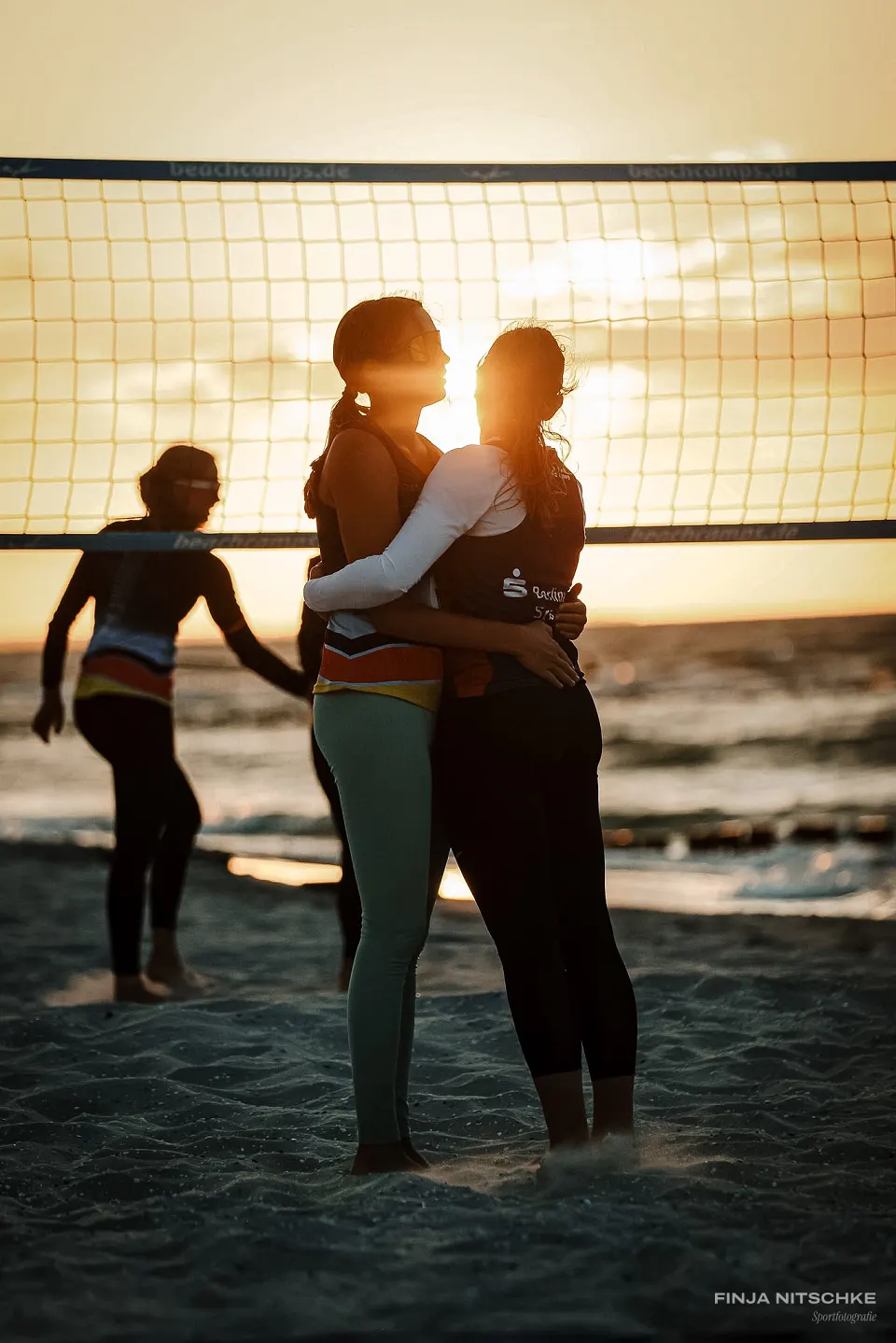 Two beach volleyball players embracing, backlit by the setting sun behind the net