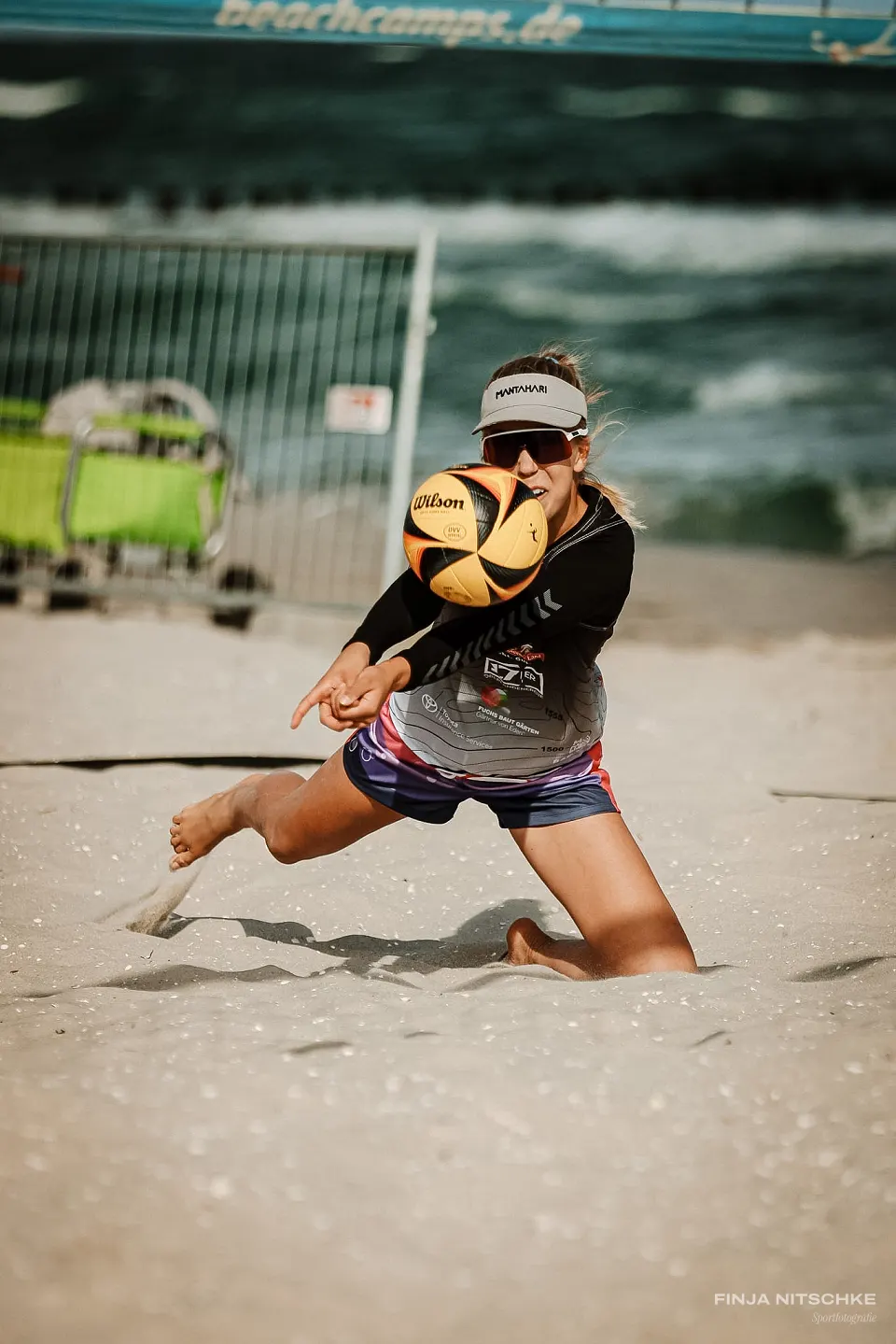 Beachvolleyballerin mit Visor baggert den Ball im Knien, Brandung im Hintergrund